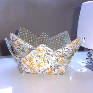May include: A fabric bowl cozy featuring a white exterior with a yellow and gray floral pattern and a gray interior lining with a yellow and white polka dot print. The bowl cozy has four folded corners and is constructed with visible stitching.