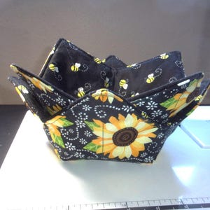 May include: Two fabric bowl cozies, one inside the other, with a sunflower and bee pattern. The outer cozy has a black background with large yellow sunflowers. The inner cozy has a black background with yellow bees.