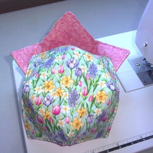May include: A fabric bowl cozy featuring a light blue background with a floral pattern of yellow daffodils, purple crocuses, and pink tulips. The interior lining is a textured pink fabric with four pointed corners extending upward from the base.