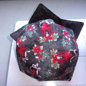 May include: A quilted fabric bowl cozy featuring a dark gray background with a repeating pattern of red lanterns, green holly leaves, red poinsettias, and pinecones. The interior of the bowl cozy is lined with a solid dark gray fabric.