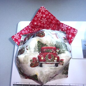 May include: A quilted fabric bowl cozy featuring a red truck with a Christmas tree and a white dog on a snowy background. The interior lining is red fabric with a white snowflake pattern. A ruler shows the base width is approximately 6 inches.