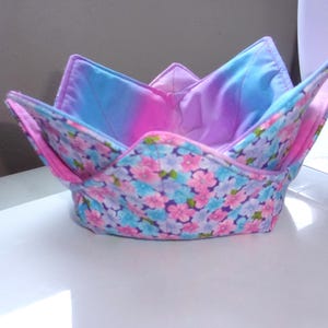 May include: A fabric bowl cozy featuring a floral pattern with pink and blue blossoms on the exterior. The interior is lined with a solid purple and blue gradient fabric. The bowl has four pointed corners and is constructed with quilted stitching.