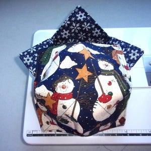 May include: A fabric bowl cozy featuring a dark blue print with snowmen, orange stars, and red scarves. The interior lining is a dark blue fabric with white snowflakes. A ruler at the base shows the item measures approximately 6 inches wide.