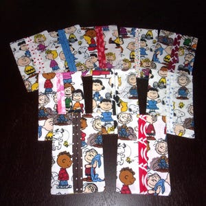 May include: A collection of handmade fabric bookmarks featuring various Peanuts characters on a white background. The bookmarks have different colored borders, including red, blue, and brown, and are arranged on a dark surface.