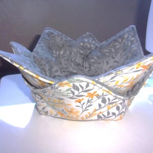 May include: A fabric bowl cozy featuring a floral pattern with yellow flowers and grey leaves on a white background. The interior is lined with a grey fabric featuring a subtle swirling pattern. The bowl has four pointed corners and stitched seams.