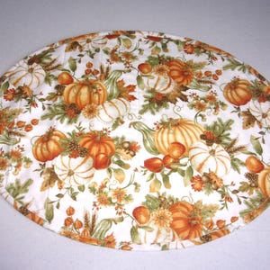 May include: An oval quilted placemat featuring a white background with a repeating pattern of orange and white pumpkins, green foliage, pinecones, and wheat stalks. The fabric has a stitched texture and a finished edge border in a matching print.
