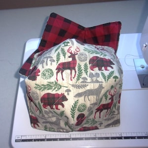 May include: A fabric bowl cozy featuring a cream background with a pattern of red and black plaid deer, bears, foxes, pine branches, and mushrooms. The interior is lined with a red and black buffalo plaid fabric. A ruler is visible at the base.