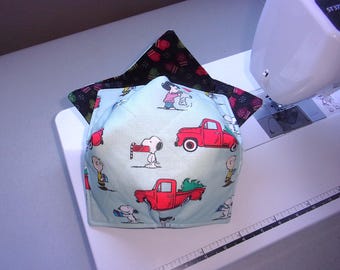 May include: A quilted fabric bowl cozy featuring a light blue exterior with a repeating pattern of Snoopy, Charlie Brown, and red pickup trucks carrying Christmas trees. The interior lining is black fabric with a colorful pattern of mittens.