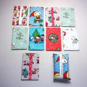 May include: Ten rectangular fabric tissue holders featuring various Peanuts character prints. Designs include Snoopy, Charlie Brown, and holiday themes with text like Be Merry & Bright and Warm & Cozy. The patterns feature red, blue, green, and white colors.