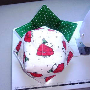 May include: A fabric bowl cozy featuring a white exterior with a repeating pattern of a cartoon dog wearing a red coat and green hat, and a green interior with white polka dots. A ruler is visible at the base showing a measurement of 12 cm.