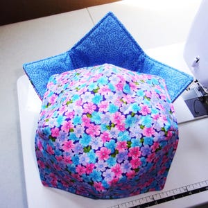 May include: A square fabric bowl cozy featuring a vibrant floral pattern with pink, light blue, and purple flowers on the exterior. The interior lining is a solid blue fabric with a textured, repeating pattern. The item is positioned on a white surface.