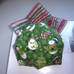 May include: A fabric bowl cozy featuring a green exterior with a pattern of snowmen, Christmas trees, and snowflakes. The interior lining displays a striped design in red, green, and white. The square-shaped item has four folded corners.