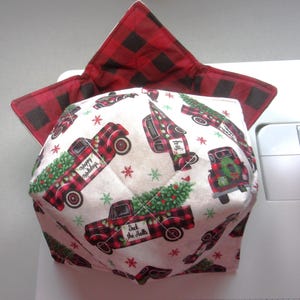 May include: A fabric bowl cozy featuring a white background with a repeating pattern of red and black plaid trucks carrying Christmas trees, accented by small snowflakes. The interior is lined with a red and black buffalo plaid fabric.