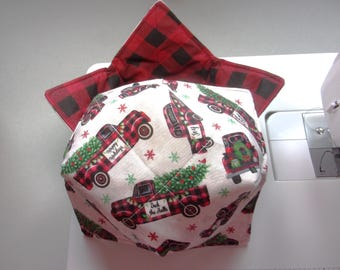 May include: A fabric bowl cozy featuring a white background with a repeating pattern of red and black buffalo plaid trucks carrying Christmas trees, accented by small snowflakes. The interior is lined with red and black buffalo plaid fabric.