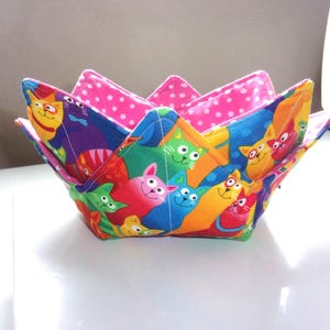 May include: A fabric bowl cozy featuring a vibrant, multi-colored cat print on the exterior with various cats in shades of blue, green, orange, red, and yellow. The interior lining is a bright pink fabric with a white polka dot pattern.