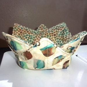 May include: A fabric bowl cozy featuring a cream-colored exterior with a pattern of stylized coffee mugs in shades of teal, brown, and tan. The interior lining displays a contrasting print of scattered brown and teal coffee beans on a tan background.