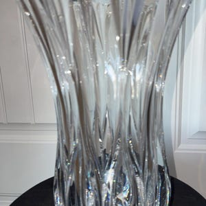 Puede incluir: Un jarrón de cristal transparente decorativo con una parte superior acampanada y un patrón vertical texturizado. El jarrón mide aproximadamente 30 cm de alto y se apoya sobre una superficie oscura. El diseño captura y refleja la luz.
