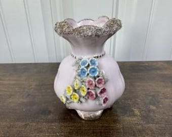 May include: A light pink ceramic vase with a ruffled rim and a silver-coloured base. The vase features a cluster of colourful, three-dimensional flowers in blue, yellow, and pink. The vase is approximately 18 cm tall.