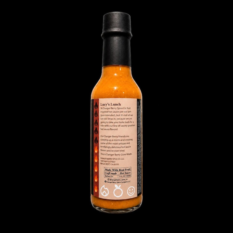 May include: A glass bottle of orange hot sauce with a black cap and a label titled Lucy's Lunch. The label features text about Danger Berry Spice Co., a column of flame icons, and the text Made With Real Fruit, Craft made Hot Sauce 5 FL OZ.