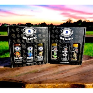 Op de afbeelding: Twee zwarte cadeaudozen met schedelontwerpen en de tekst "Core Punk Hot Sauces" op de voorkant. Elke doos bevat drie kleine glazen flesjes met hete saus met kleurrijke etiketten met cartoonfiguren en namen zoals "Pinco Pingo", "Blue-Fi", "Killer Kai", "Core Punk", "Core Punk" en "Haizenz",