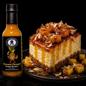 K&ouml;nnte beinhalten: Eine Flasche Danger Berry Spice Co. Pyromania Pineapple scharfe So&szlig;e neben einem quadratischen St&uuml;ck K&auml;sekuchen, belegt mit karamellisierten Ananasringen, Kokosflocken und einer goldenen Glasur, vor einem schwarzen Hintergrund.