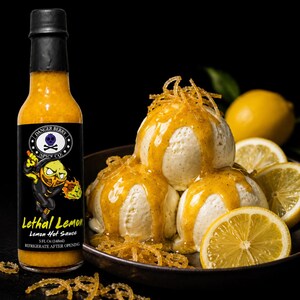 Op de afbeelding: Een glazen fles Danger Berry Spice Co. Lethal Lemon hete saus met een zwarte dop en een etiket met een geel citroenhoofd-figuur. Daarnaast drie bollen vanille-ijs overgoten met gele hete saus en citroenrasp.