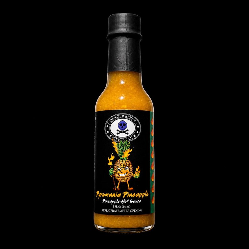 K&ouml;nnte beinhalten: Eine klare Glasflasche mit Pyromania Pineapple scharfer So&szlig;e und schwarzem Verschluss. Das Etikett zeigt eine animierte Ananasfigur mit Flammen, ein Totenkopf-Logo und den Text Danger Berry Spice Co. Die Flasche enth&auml;lt 148 ml der So&szlig;e.
