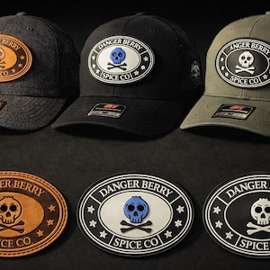 May include: Three baseball caps in charcoal, black, and olive green, each featuring an oval patch with a skull and crossbones logo. The text reads DANGER BERRY SPICE CO. Three matching oval patches are displayed below the caps, showing variations in color and material texture, including tan leather, white with a blue skull, and black with a white skull.
