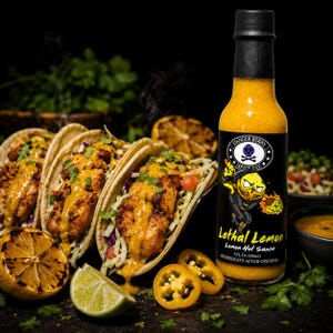 Op de afbeelding: Een glazen fles Danger Berry Spice Co. Lethal Lemon hete saus met een zwarte dop en een etiket met een gele stripfiguur. De fles bevat 148 ml gele saus en staat naast drie vistaco's en partjes limoen op een donkere ondergrond.