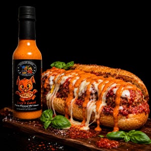 May include: A bottle of Danger Berry Spice Co. Lucy's Lunch pizza flavored hot sauce next to a meatball sub on a toasted baguette, topped with melted white cheese, red tomato sauce, and drizzled with orange hot sauce, garnished with basil.