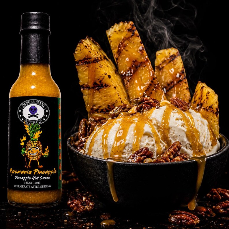 K&ouml;nnte beinhalten: Eine Flasche Danger Berry Spice Co. Pyromania Pineapple scharfe So&szlig;e neben einer schwarzen Sch&uuml;ssel mit Vanilleeis, garniert mit gegrillten Ananasscheiben, Pekann&uuml;ssen und einem goldenen Sirup-Guss, mit sichtbarem Dampf &uuml;ber der Sch&uuml;ssel.