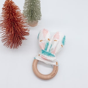 May include: A white and pink fabric baby teether with a wooden ring. The fabric has a teal and gold pattern. The ring has the words "Burton Baby" engraved on it.