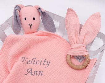 Può includere: Una copertina per neonati in mussola rosa con un sonaglio a forma di coniglio e un mordedor di legno. La copertina è personalizzata con il nome "Felicity Ann".