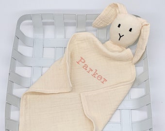 Può includere: Una copertina per neonati in mussola color crema con una testa di coniglio attaccata. La copertina è personalizzata con il nome "Parker" ricamato in filo rosa.