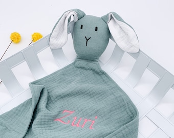 Può includere: Una morbida copertina per neonati in mussola verde con una testa di coniglio cucita. Il coniglio ha orecchie bianche e un viso ricamato in nero. La copertina è personalizzata con il nome "Zuri" in ricamo rosa.