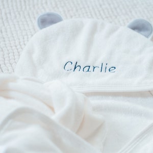 May include: White hooded towel with gray ears and the name "Charlie" embroidered in blue thread.