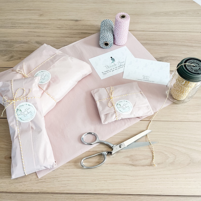 May include: Three small packages wrapped in pink tissue paper with yellow twine and a round sticker that says "The Button Baby" are on a wooden surface.  A pair of silver scissors, two spools of twine, and a thank you card are also on the surface.