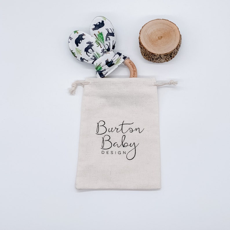May include: A white muslin drawstring bag with the text "Burton Baby Design" printed on it. The bag contains a wooden teether with a moose and bear print fabric covering. The teether is on a white background.