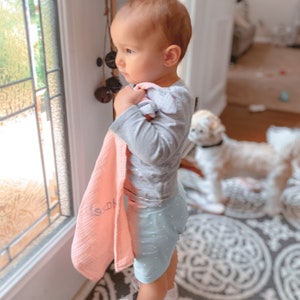 May include: A young child wearing a grey long-sleeved shirt and blue polka dot shorts holds a pink muslin swaddle blanket with the word "Love" embroidered on it. The child is looking out a window.