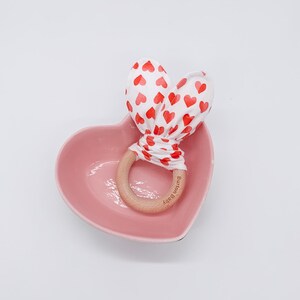 Op de afbeelding: Een houten bijtring met de tekst "Burton Baby" erop gegraveerd. De ring is vastgebonden met een witte stof met een rood hartpatroon. De ring ligt in een roze hartvormige schaal.
