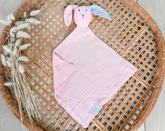 Può includere: Una coperta di sicurezza rosa per bambini con testa e orecchie di coniglio. La testa del coniglio è rosa con orecchie blu. La coperta quadrata è in tessuto morbido e strutturato. Su un vassoio di vimini intrecciato.