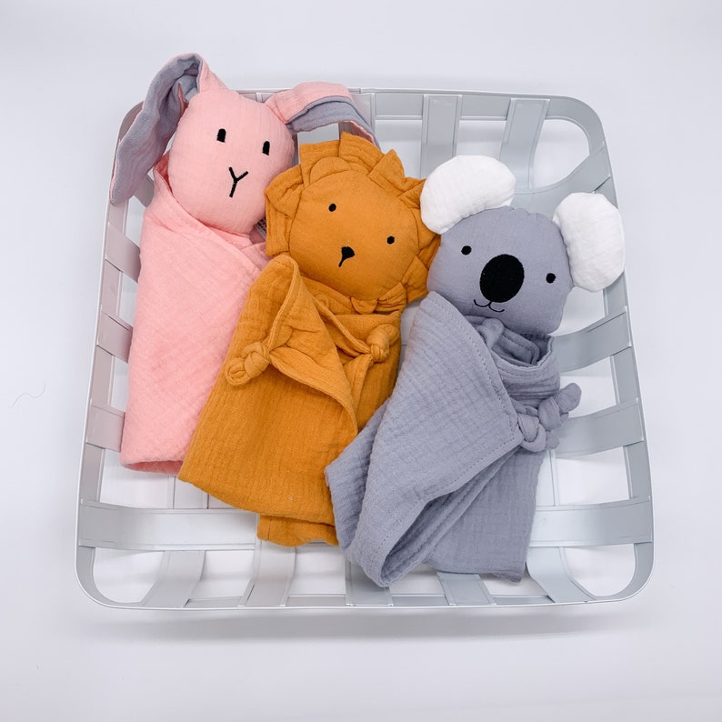 May include: Three plush toys, a pink bunny, an orange lion, and a gray koala, are wrapped in soft muslin blankets. The toys are arranged in a white wicker basket.