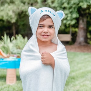 May include: A white hooded towel with blue ears and the name "Asher" embroidered on the hood. A young child is wearing the towel and smiling at the camera.