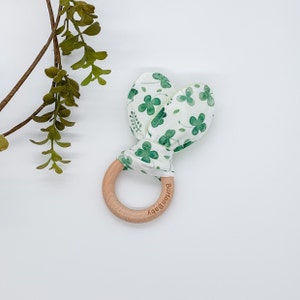 May include: A white and green fabric baby teether with a clover pattern. The teether is attached to a wooden ring with the word "Burtonbaby" engraved on it.
