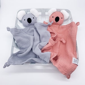 May include: Two soft, plush koala baby security blankets. One is gray and the other is pink. Both have black eyes and a black nose. The blankets are made of a soft, textured fabric.