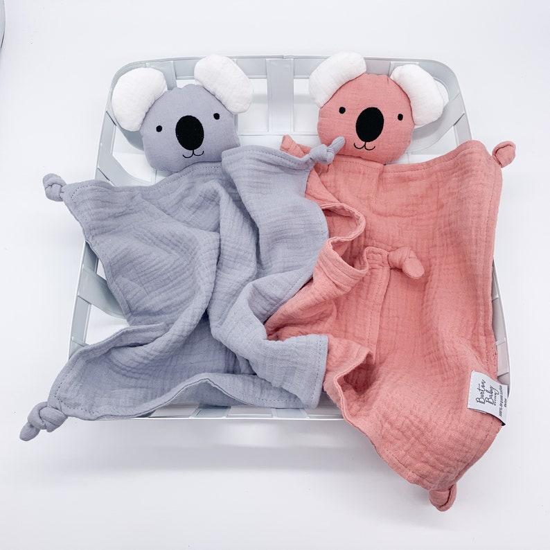 May include: Two soft, plush koala baby security blankets. One is gray and the other is pink. Both have black eyes and a black nose. The blankets are made of a soft, textured fabric.