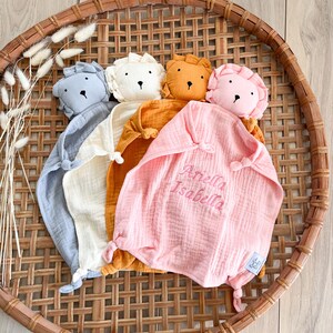May include: Four plush lion-shaped baby security blankets in different colors. The blankets are made of soft, textured fabric and have a knotted corner. The pink blanket has the name "Ariella Isabella" embroidered on it.