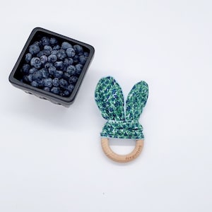 May include: A wooden teether with blue and green fabric bunny ears. The fabric has a blueberry print. The teether is labeled "buttonbaby".