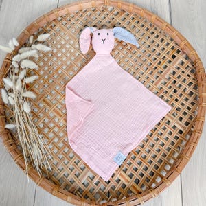 May include: Pink muslin baby security blanket with a bunny head attached. The bunny has gray ears and a pink face with embroidered eyes and a nose.