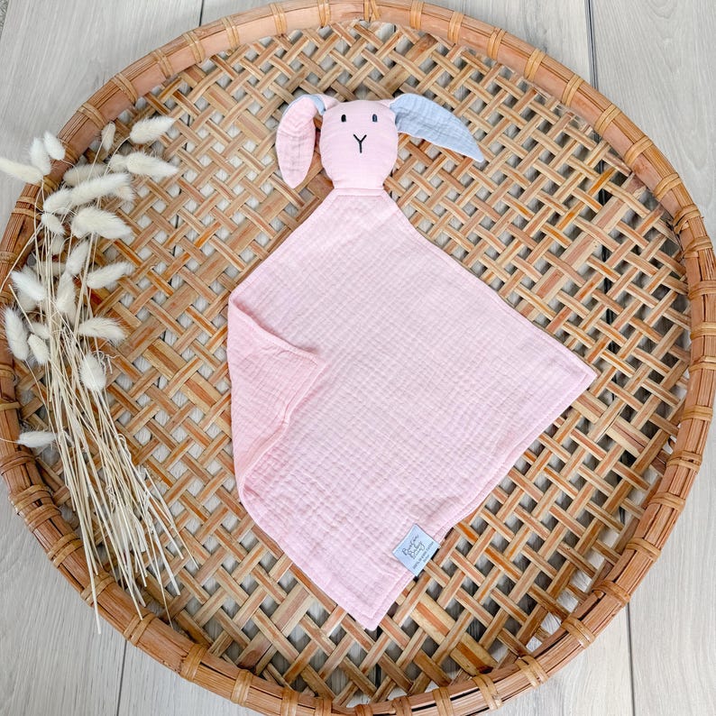 May include: Pink muslin baby security blanket with a bunny head attached. The bunny has gray ears and a pink face with embroidered eyes and a nose.